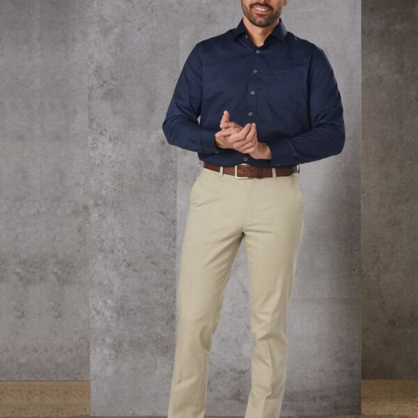 A man in a navy shirt and beige pants stands against a gray background, smiling.