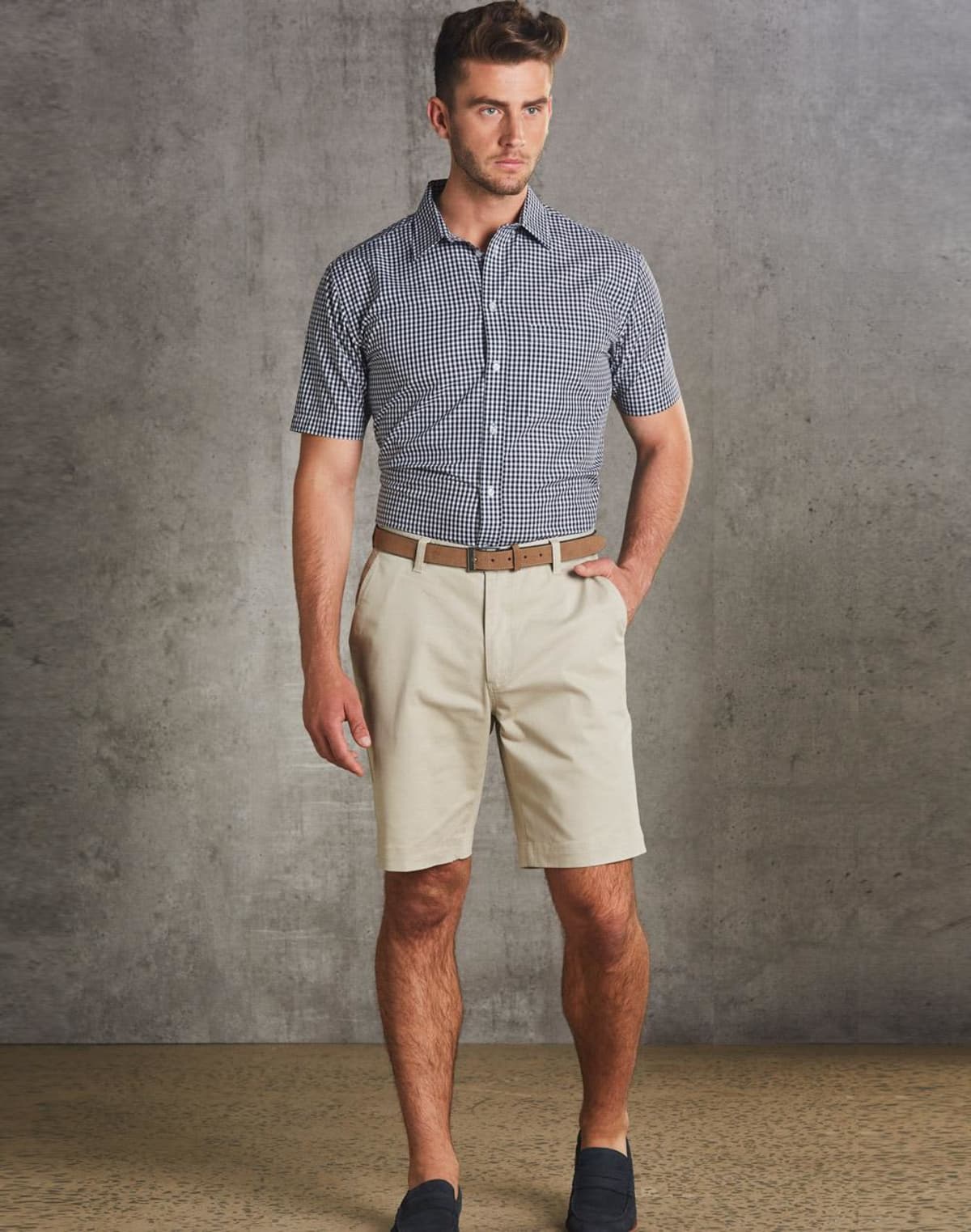 Man in checkered shirt, beige shorts, and loafers standing against a gray background.