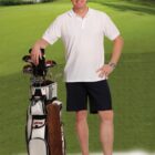 Man in white polo and shorts stands with golf bag on sunny golf course.