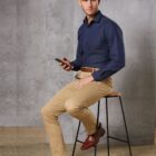 Man in navy shirt and khaki pants sits on stool, holding phone, with textured gray background.