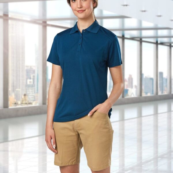 Person in a blue polo and khaki shorts stands in a modern, glass-walled room with a city view.