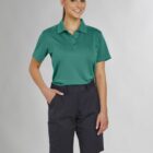 Person in green polo and dark shorts standing, smiling with one hand in pocket.