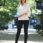 Woman in a white blouse and black pants stands outdoors on a tree-lined pathway.