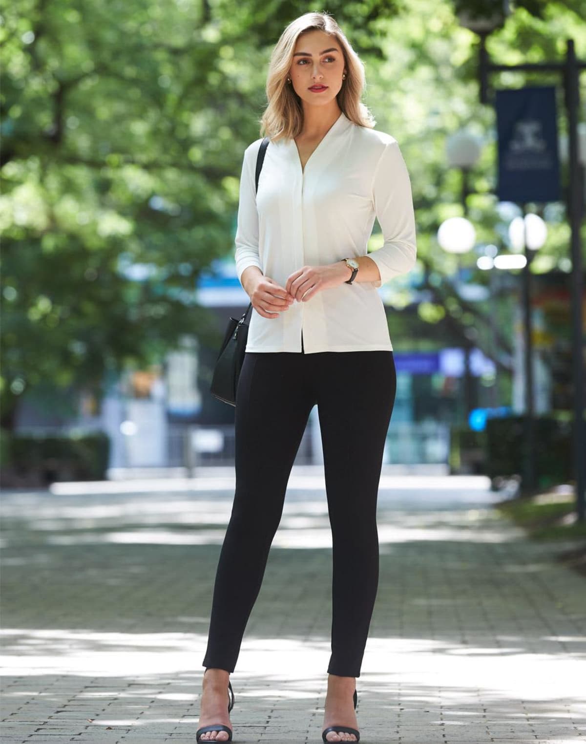 Woman in a white blouse and black pants stands outdoors on a tree-lined pathway.