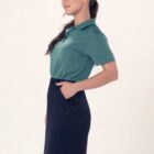 Person in a green polo and black skirt stands in profile with hand in pocket.