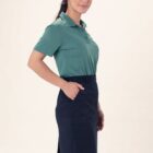 Person in green shirt and black skirt standing sideways with hands in pockets.