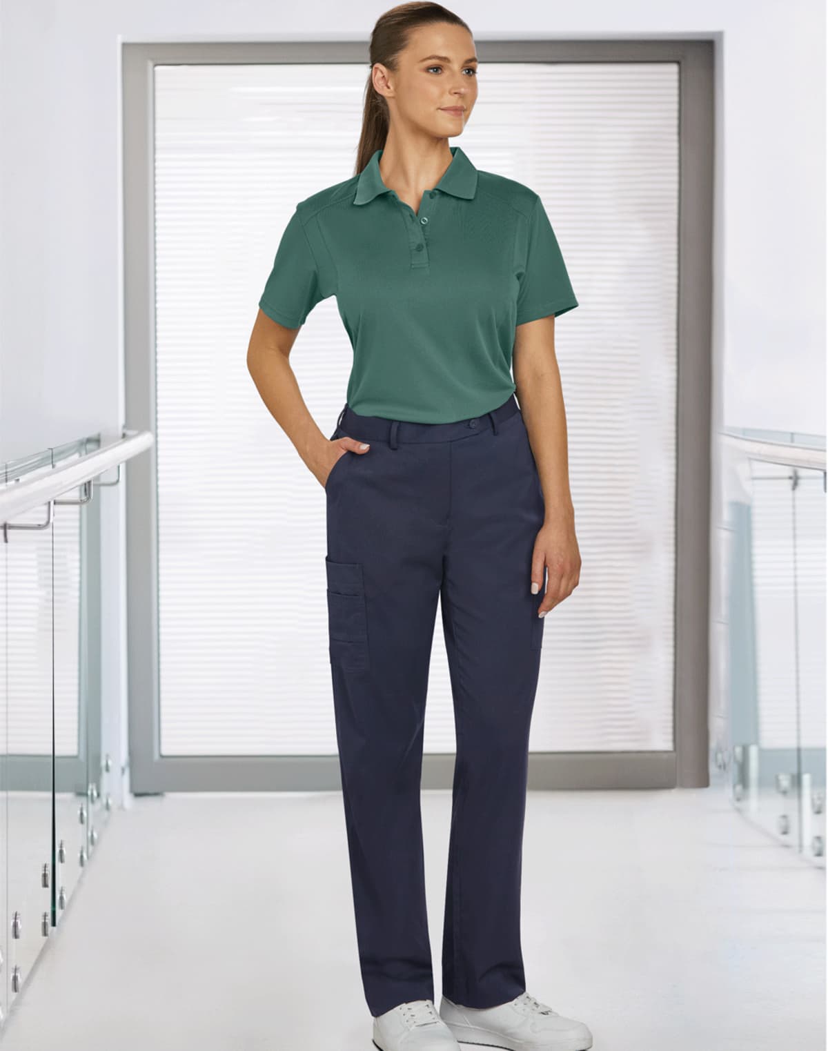 Woman in green polo and navy pants stands in a bright, modern hallway, hand in her pocket.