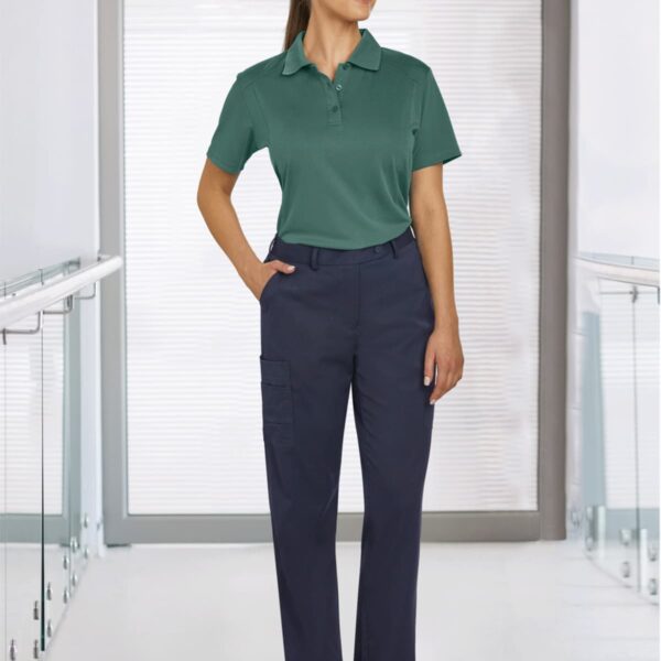 Person in a green polo, navy pants, and white shoes standing in a hallway with large windows.