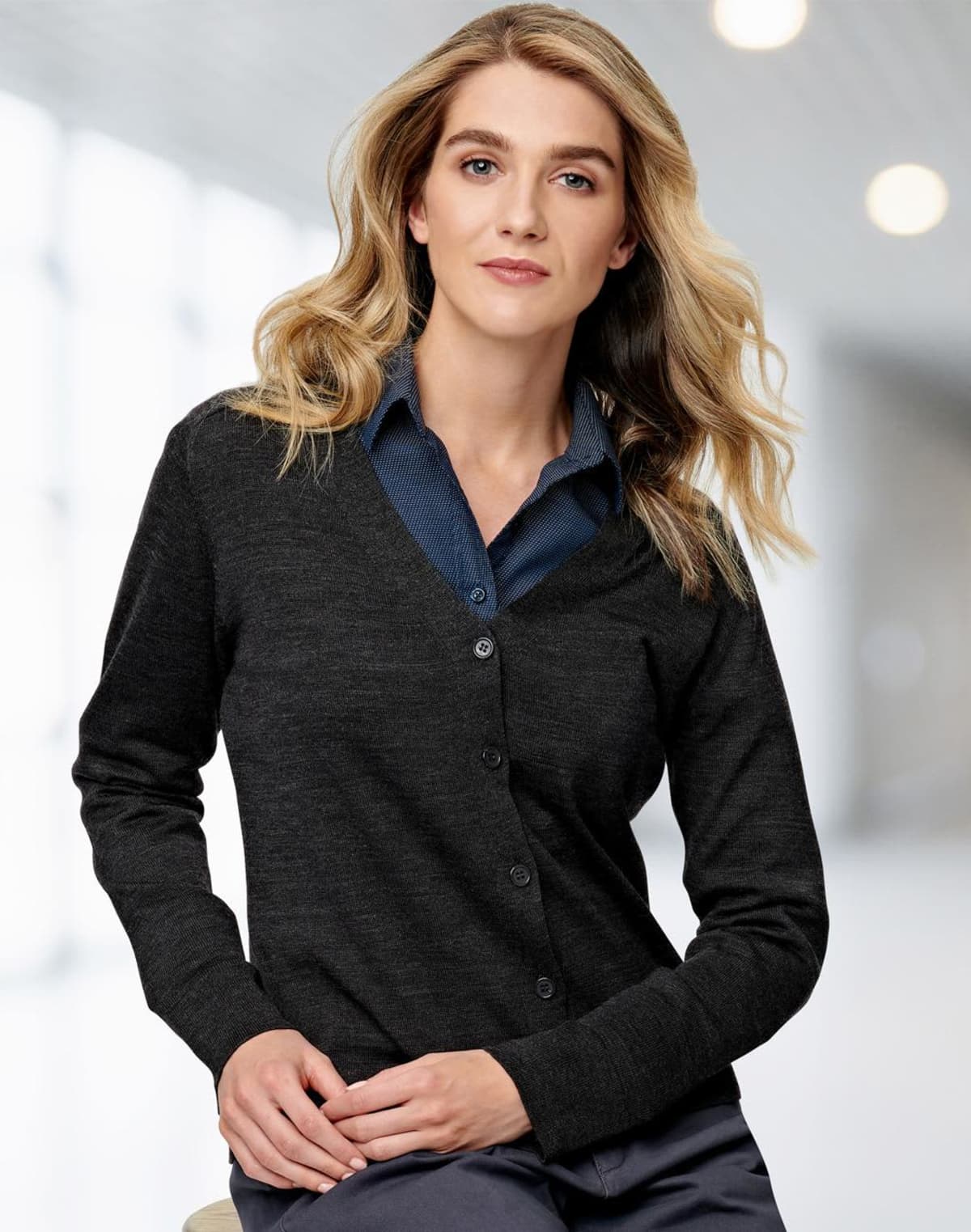 Woman in a dark cardigan and blue shirt, standing indoors with blurred background.