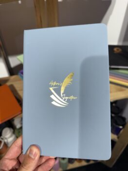 A hand holds a blue notebook with Branded Office Supplies and a gold feather on the cover.