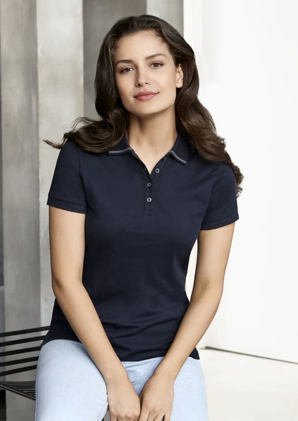 Woman with long brown hair wearing a navy polo shirt and light jeans, sitting indoors.