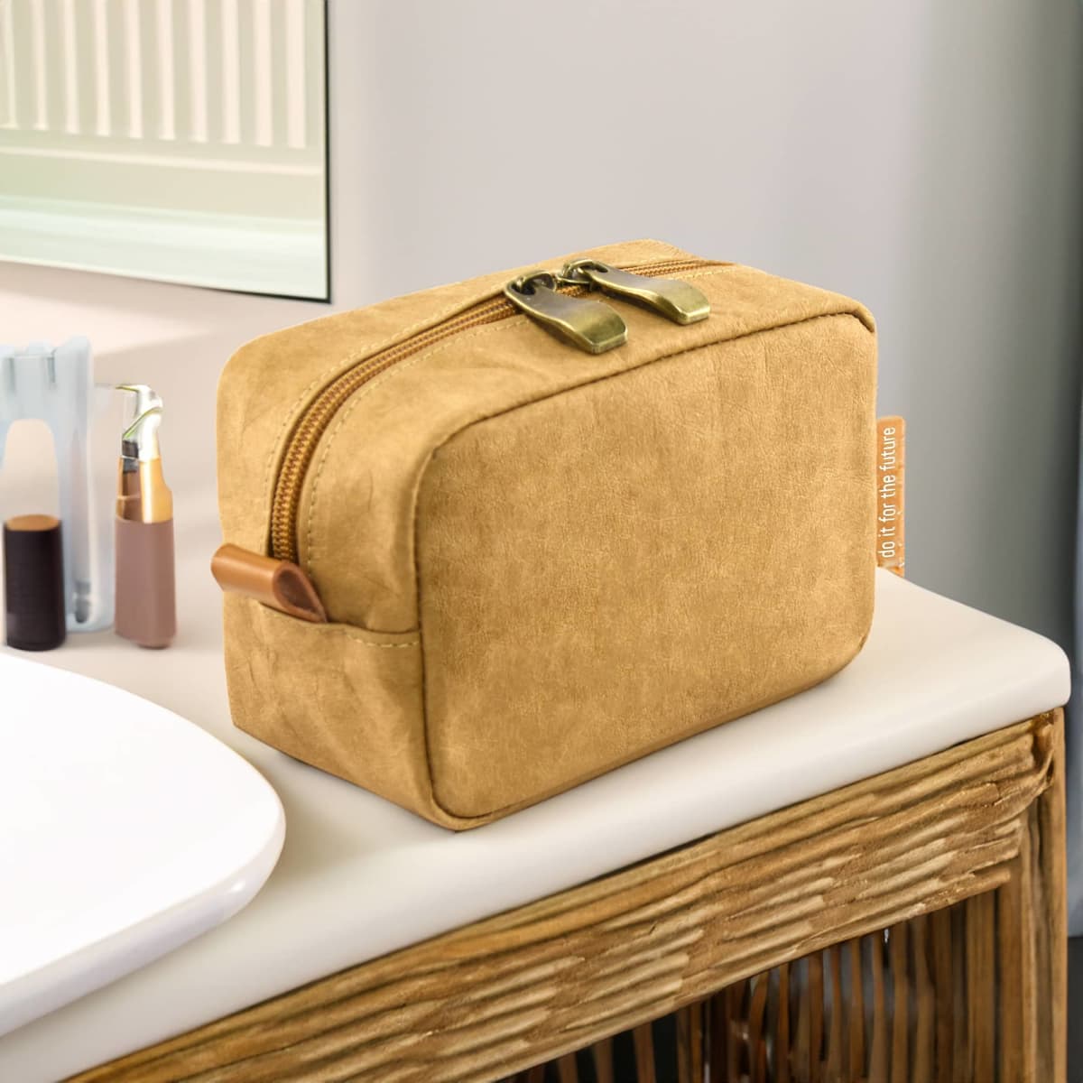 Tan toiletry bag with zipper on a wicker surface beside a bathroom sink and toiletries.