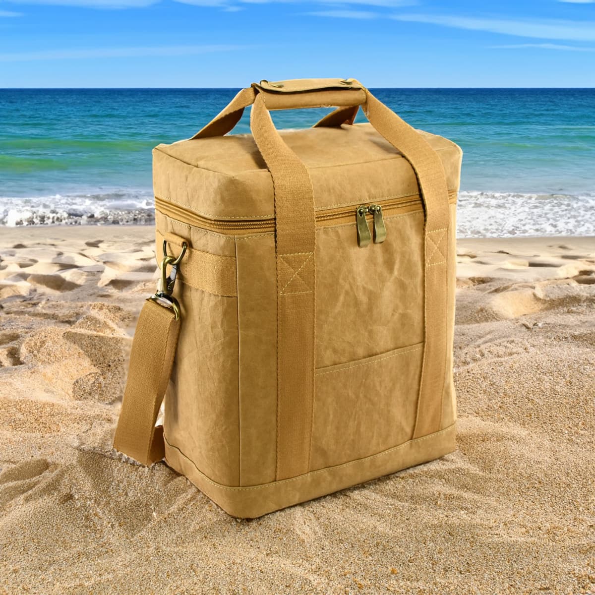 Tan insulated cooler bag on sandy beach with ocean waves and blue sky in the background.