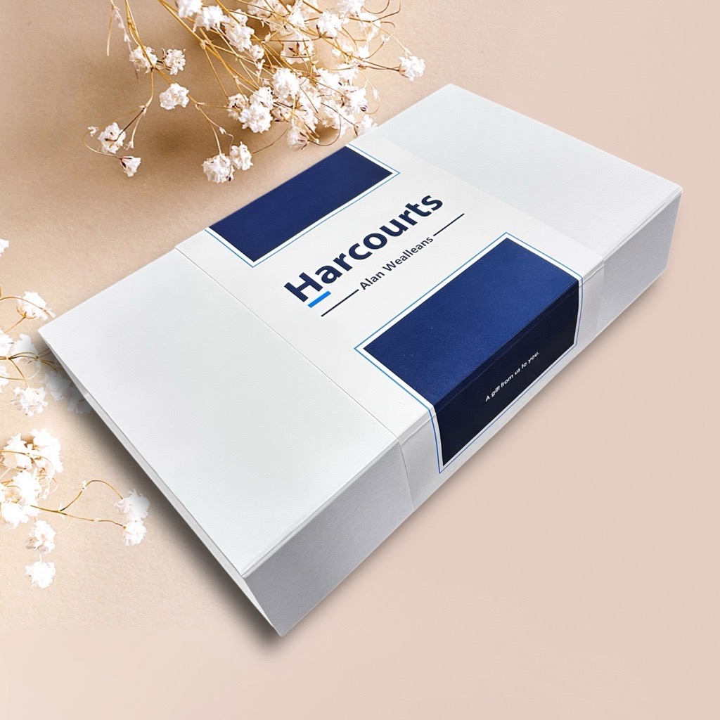Gift boxes with Harcourts logo, blue accents, on a beige surface with white flowers.