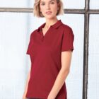 Woman in a red polo shirt and jeans standing indoors by a window, facing the camera.