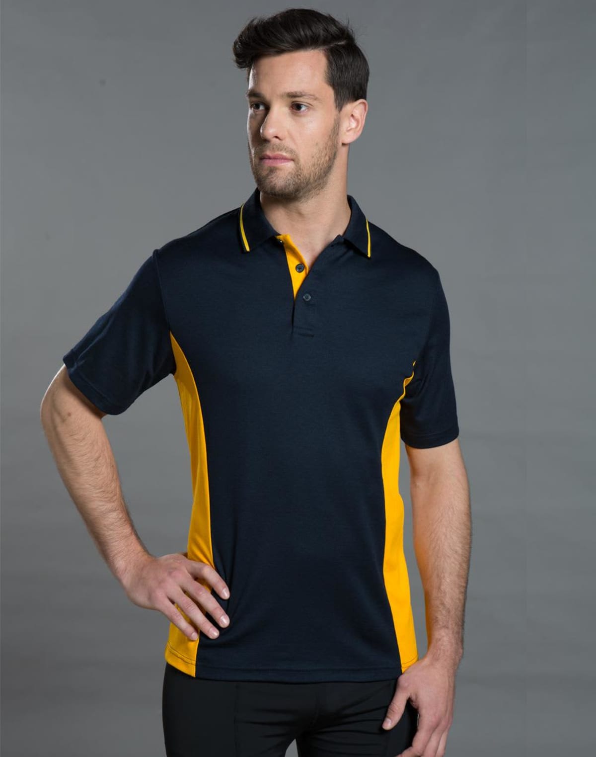 Man wearing a navy and yellow polo shirt with one hand on hip, against a gray background.
