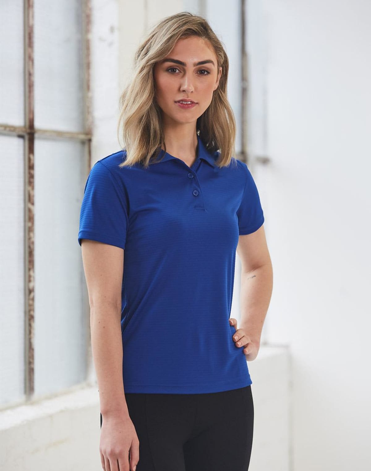Person standing in a blue polo shirt and black pants near a window.