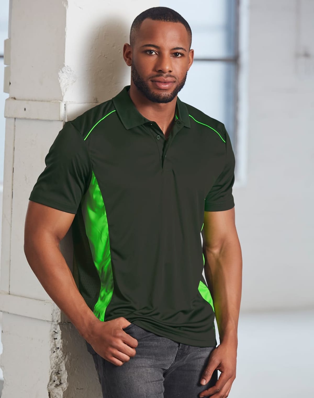 Man in a green and black polo shirt leans against a wall in a bright room.