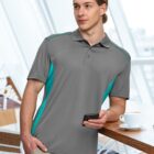 Person in a gray polo shirt holding a phone, standing by a table with a cup.