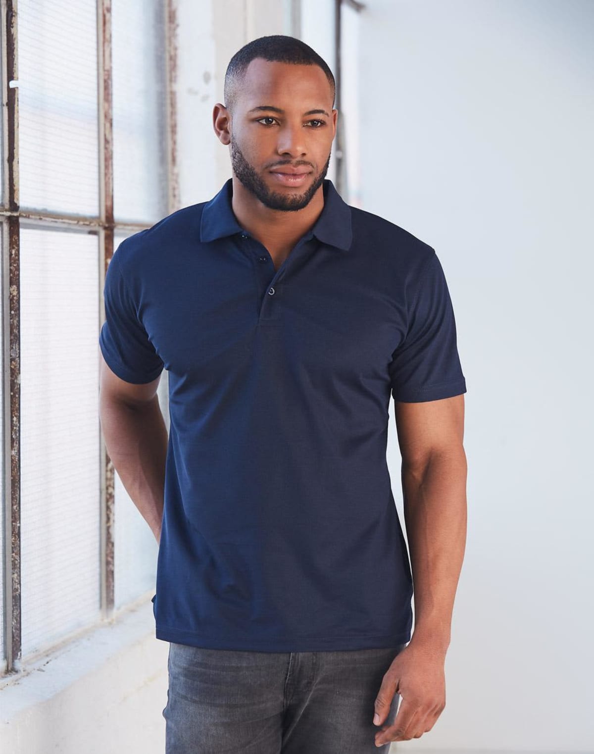 Person in a dark polo shirt stands near a window, looking thoughtful.