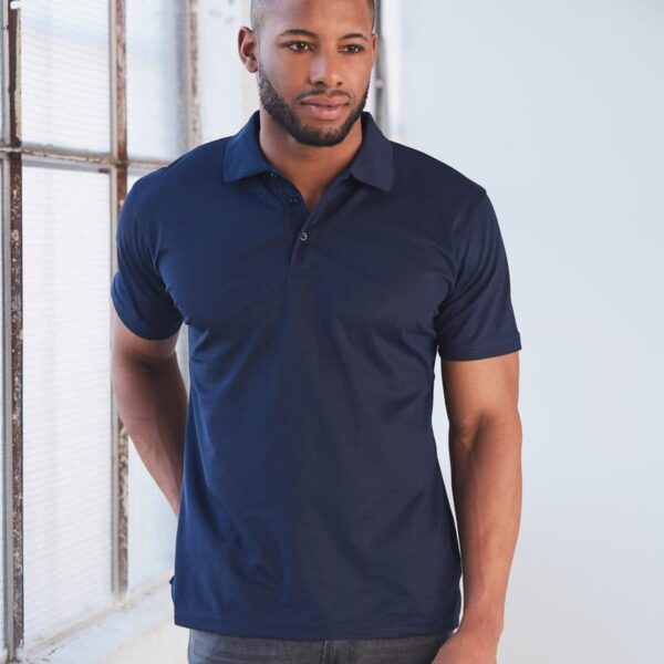 Man in a navy polo shirt stands by a window, looking into the distance.