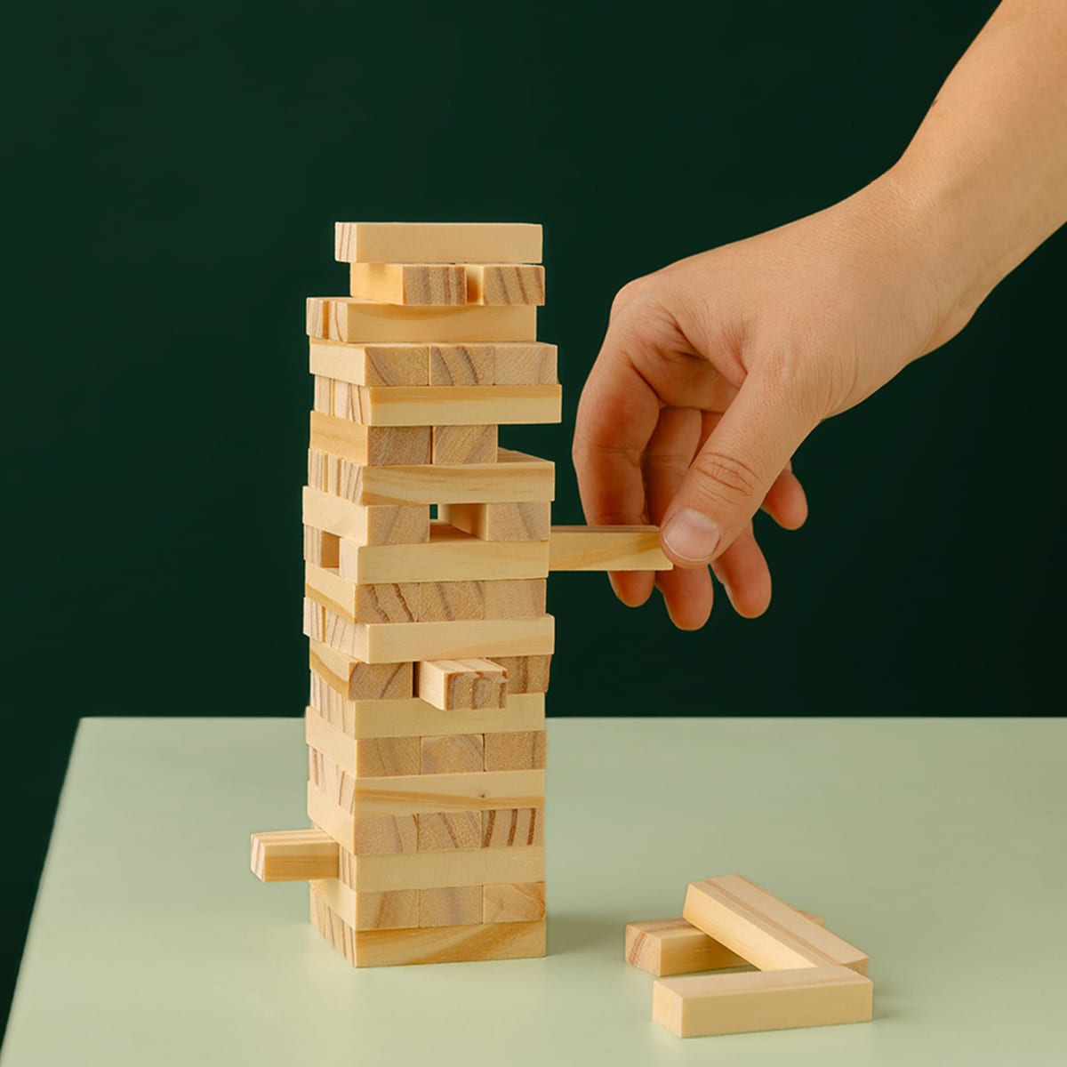 A hand pulls a block from a stacked wooden Jenga tower on a green surface.