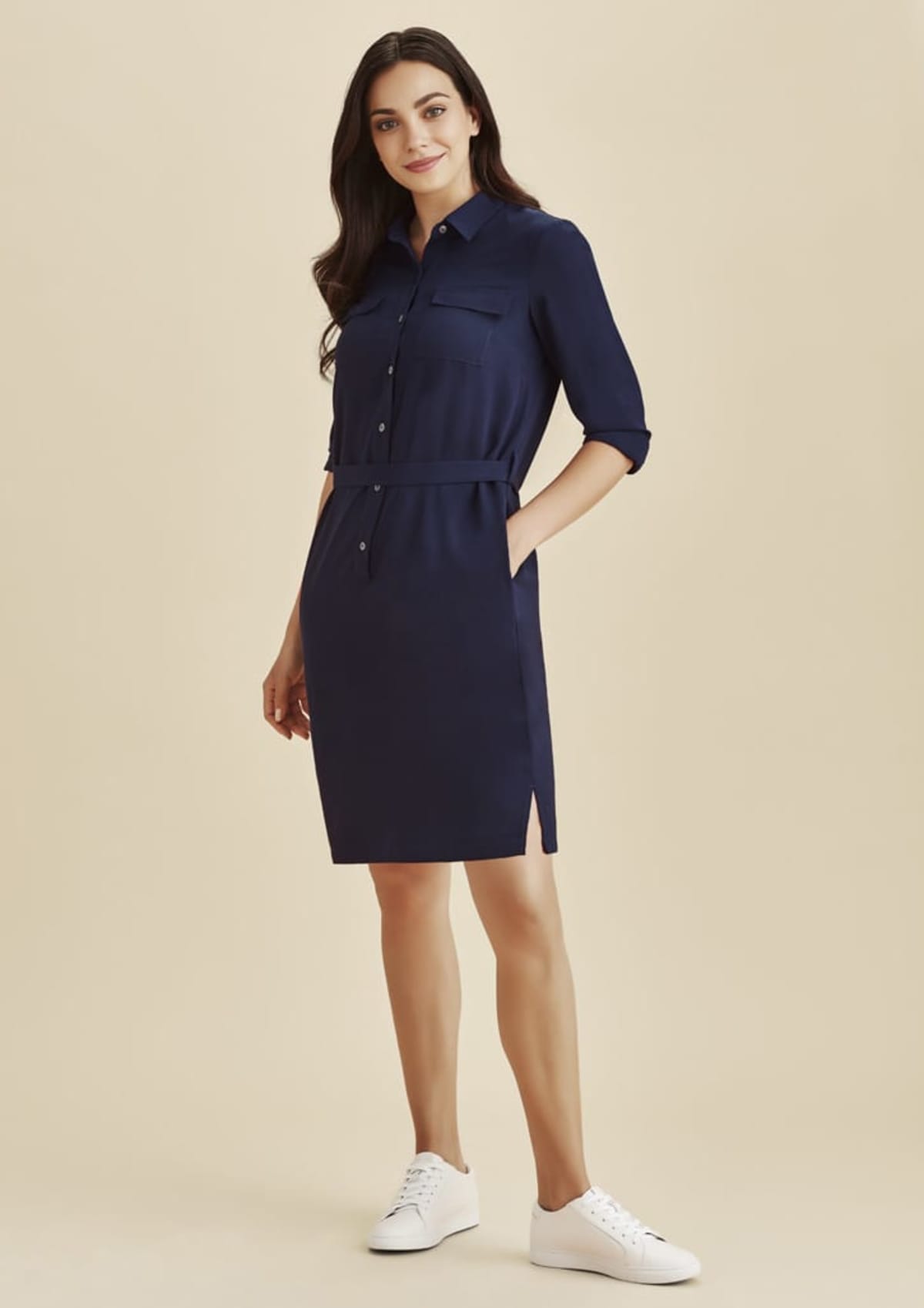 Woman in a navy shirt dress and white sneakers, standing against a beige background.