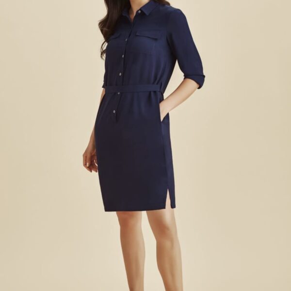 Woman in a navy shirt dress and white sneakers standing against a beige background.