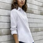 Woman in a white button-up shirt and jeans leans against a gray brick wall.