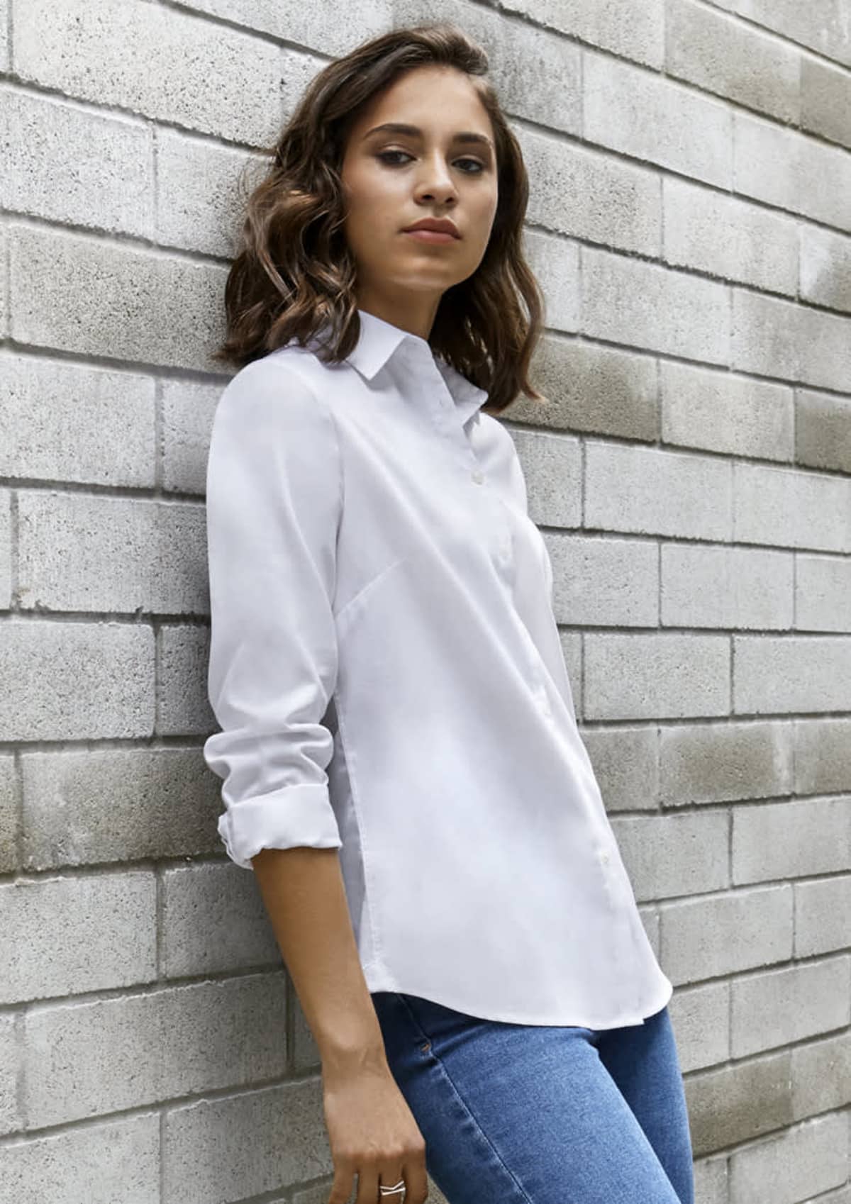 Woman in a white button-up shirt and jeans leans against a gray brick wall.