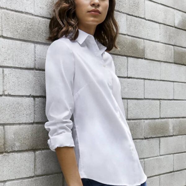 Woman in a white shirt and jeans leaning against a light gray brick wall.