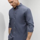 Man wearing a long-sleeve, button-up gray shirt with sleeves rolled up, looking down.