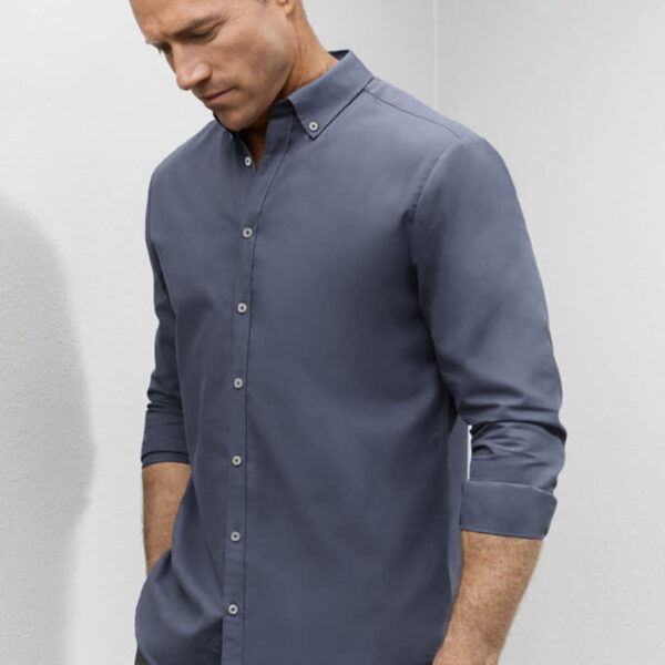 Man wearing a fitted, long-sleeve, button-up gray shirt, standing against a light wall.