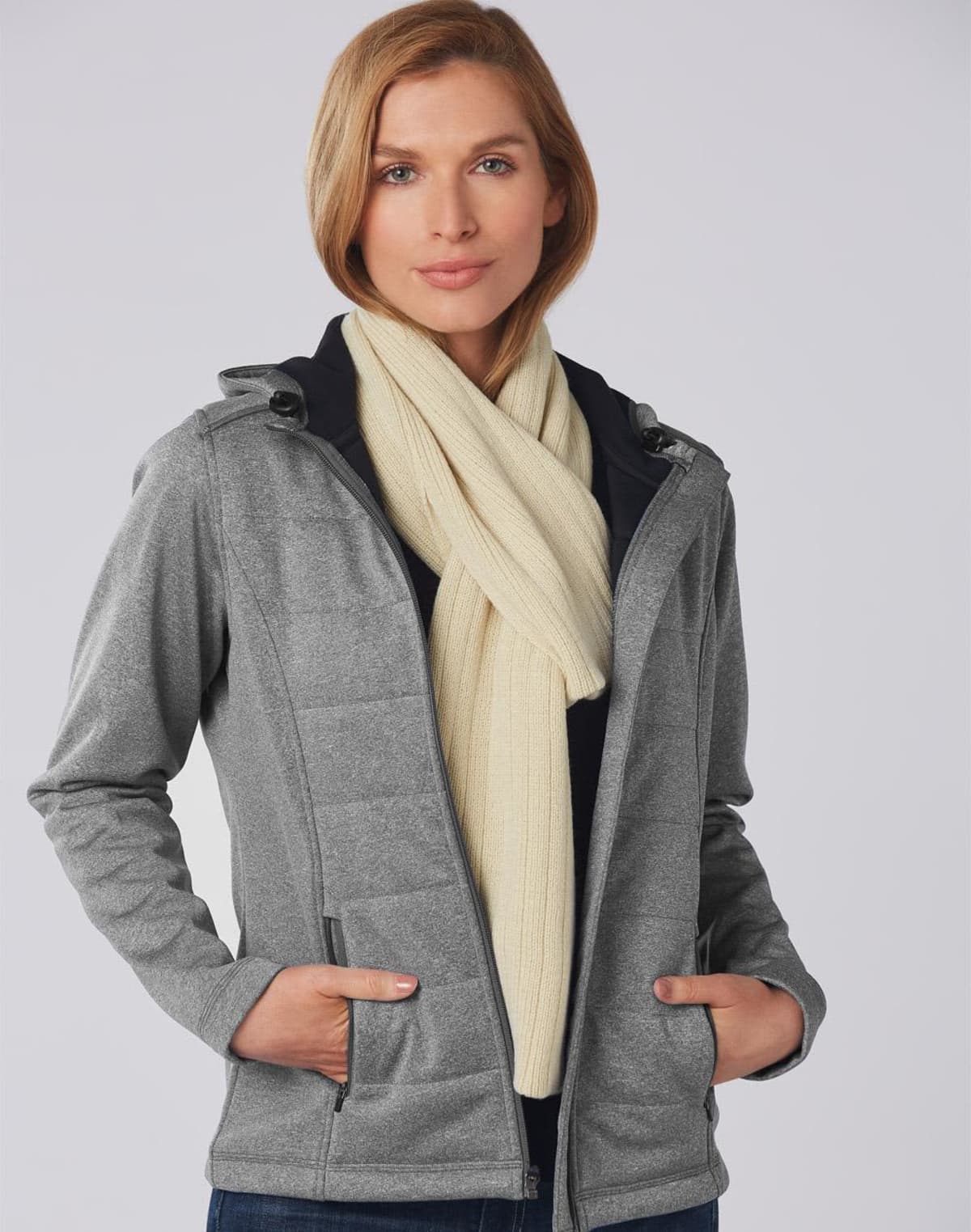 Person in a gray jacket and cream scarf, hands in pockets, standing against a plain background.