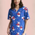 Smiling person wearing a blue Santa-themed scrub top on a pink background.