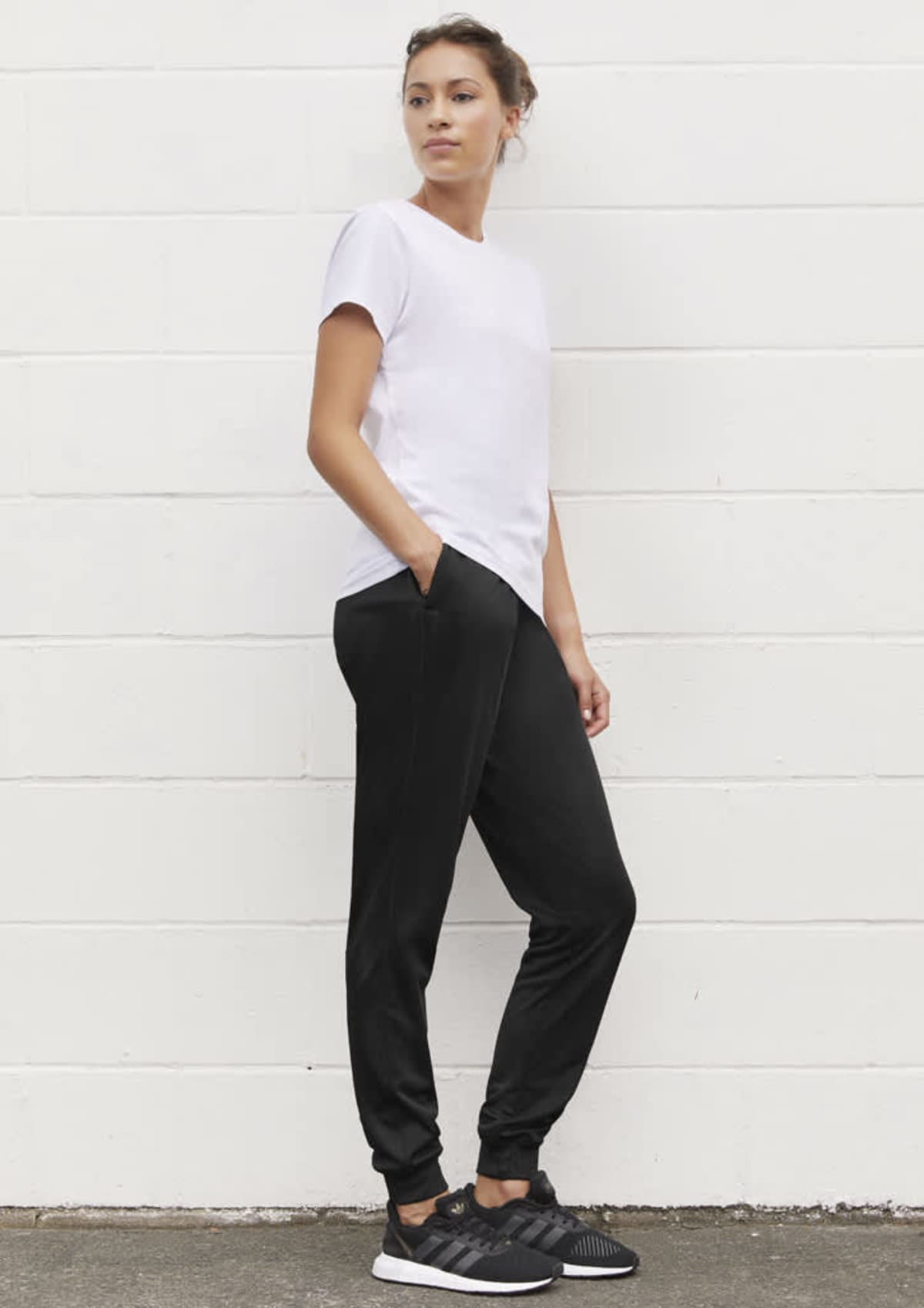 Person in a white T-shirt and black joggers, standing against a white brick wall.