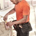 Person in an orange shirt measuring wood with a tape measure and pencil.