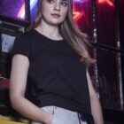 Woman in a gray beanie and black t-shirt stands by a window with neon lights at night.