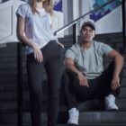A woman stands and a man sits on stairs in casual wear, with a mural in the background.