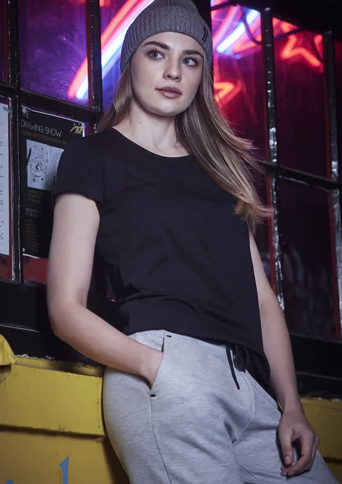 Woman in a gray beanie, black t-shirt, and gray pants stands by a window with neon lights.