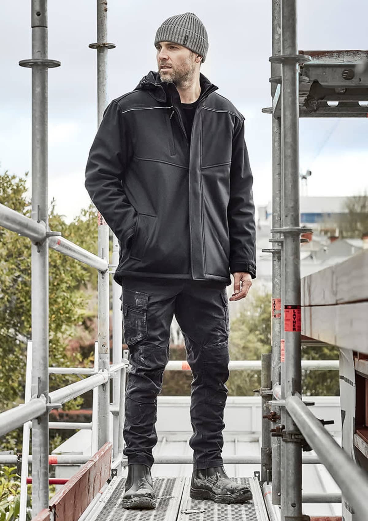 Man in black jacket and beanie standing on scaffolding, looking into the distance.