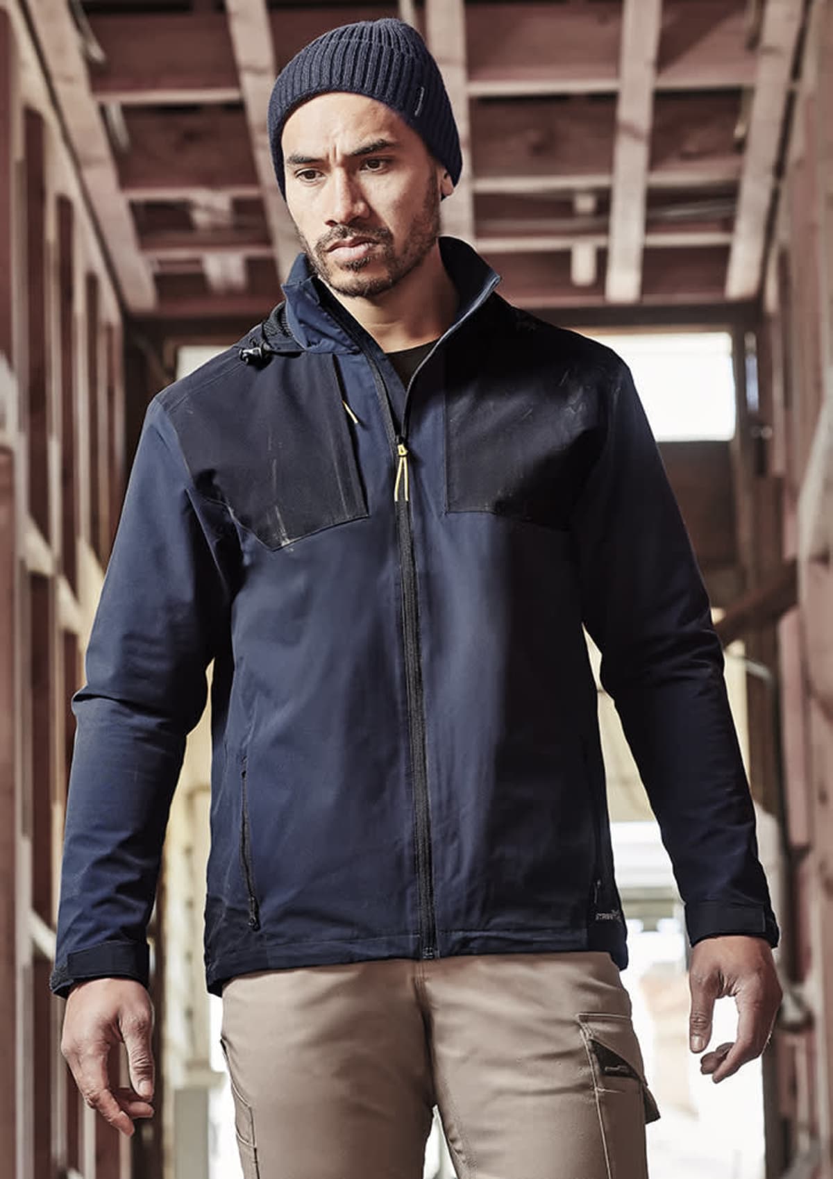 Man in a navy jacket and beanie stands in a wooden structure, looking to the side.