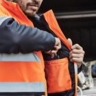 Man wearing an orange vest checks pocket contents outdoors.