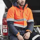 Person in an orange safety jacket sits on a vehicle, wearing a beanie.