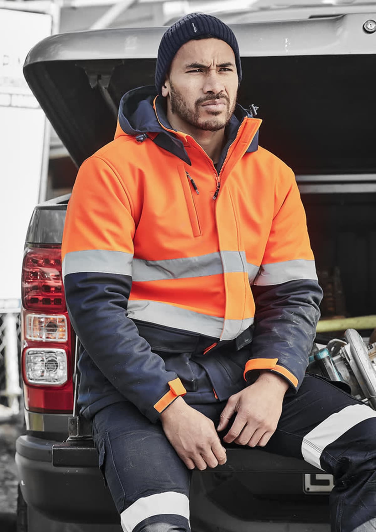 Person in an orange safety jacket sits on a vehicle, wearing a beanie.