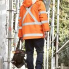 Person in orange safety jacket and jeans walking at a construction site, carrying a tool bag.