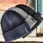 Three folded knit beanies in navy, gray, and black on a wooden surface.