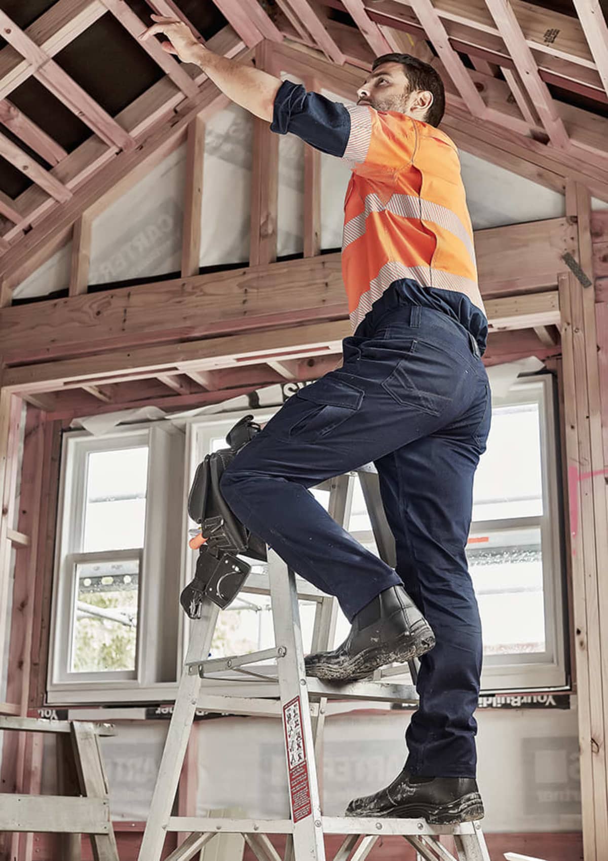 Worker in safety gear stands on ladder, reaching up inside a house under construction.