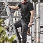 Man in workwear standing on a ladder at a construction site, holding a tool.
