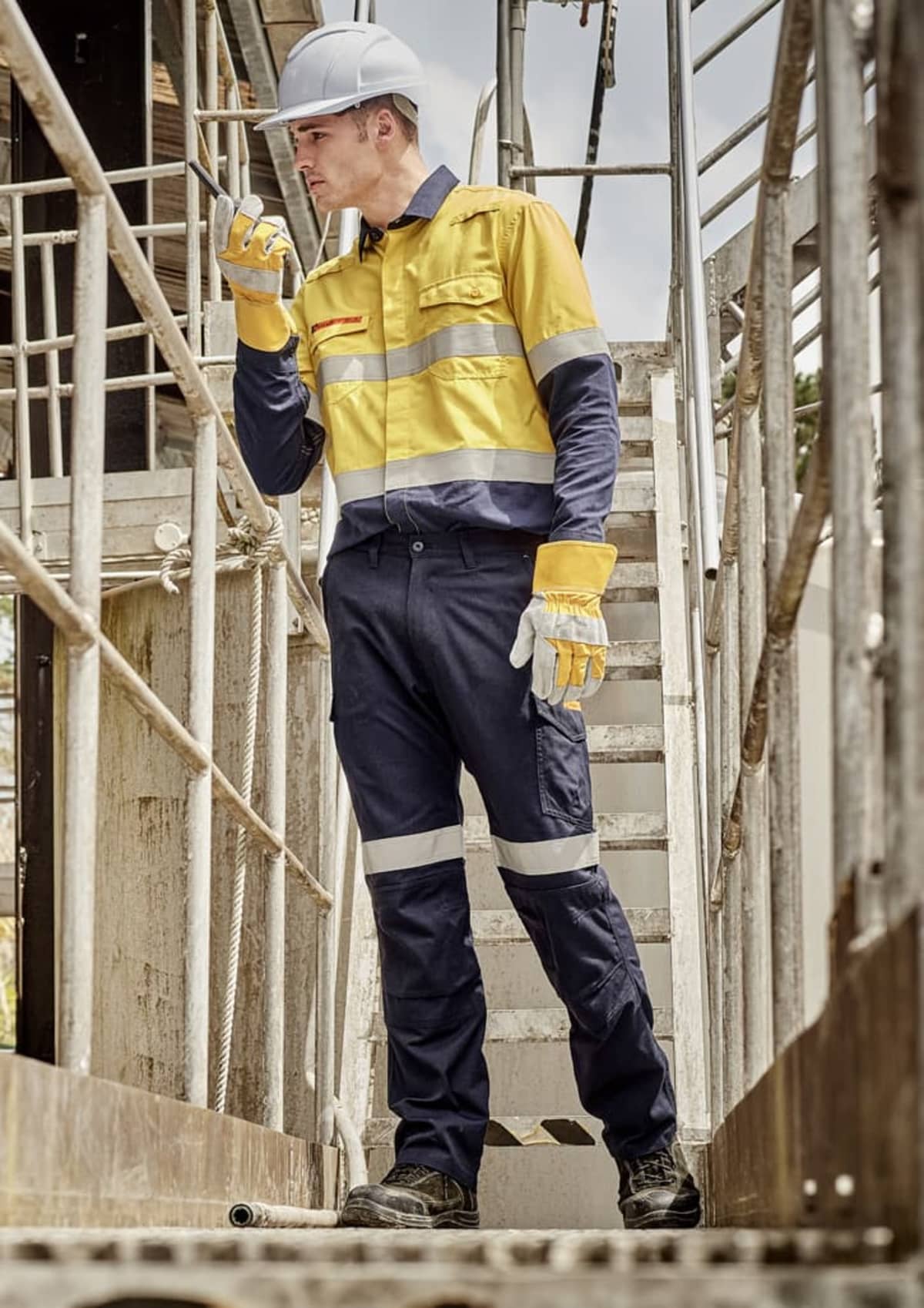 Worker in yellow and blue safety gear, holding a radio, stands on an industrial walkway.